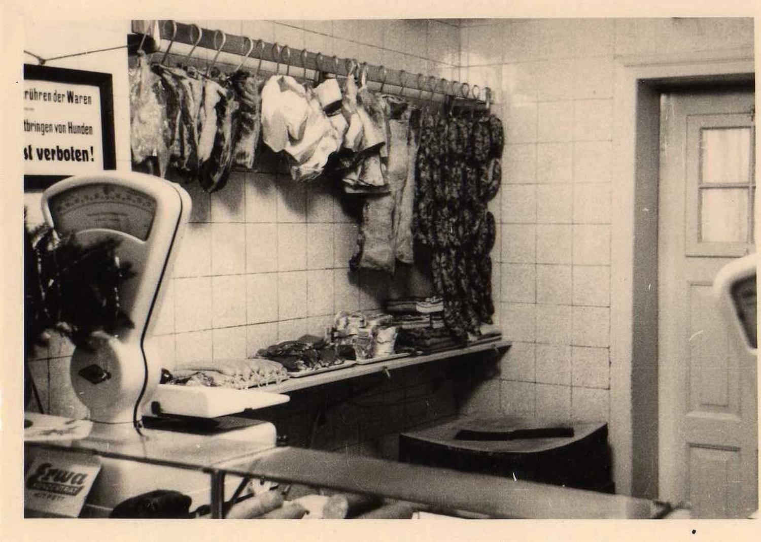 Hecht – Fleischerei – Das Bild zeigt das Innere einer traditionellen europäischen Metzgerei, wahrscheinlich aus dem frühen bis mittleren 20. Jahrhundert, mit Fleisch, das an Haken hinter einer Theke hängt.